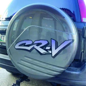 May include: A grey spare wheel cover featuring the letters "CR-V" in white and purple. The cover is mounted on the rear of a dark grey SUV. The vehicle has a black trailer hitch and a chrome exhaust pipe.