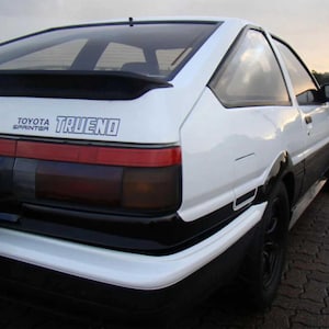 May include: White Toyota Sprinter Trueno coupe with black detailing. The car has a rear spoiler, black wheels, and a red and black taillight design. The text "TOYOTA SPRINTER TRUENO" is displayed on the rear.