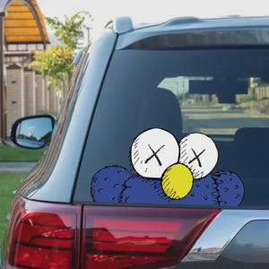 May include: A blue and white car window sticker depicting a cartoon character with white eyes, black 'X' marks, and a yellow nose. The sticker is applied to the rear window of a grey car, with the taillight visible.