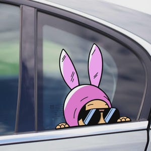 May include: A car window decal featuring a cartoon character wearing a pink bunny hat and sunglasses. The character is peeking out from the car window. The decal is a fun and playful accessory.
