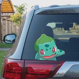 Puede incluir: Una calcomanía de Bulbasaur turquesa está adherida a la ventana trasera de un coche gris. El personaje de dibujos animados tiene ojos rojos, una boca sonriente y un bulbo verde en la espalda. El piloto trasero del coche es visible.