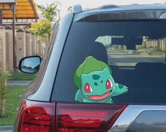 May include: A teal Bulbasaur decal is affixed to the rear window of a gray car. The cartoon character has red eyes, a smiling mouth, and a green bulb on its back. The car's taillight is visible.