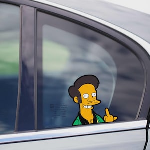 May include: A cartoon sticker of a man with dark hair, yellow skin, and a green shirt giving the middle finger. The sticker is affixed to a car window, with the car's exterior visible in the background.