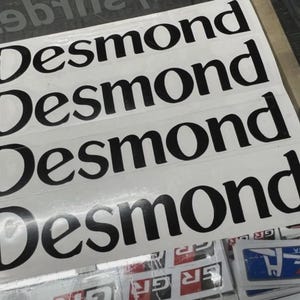 May include: A stack of white stickers with the name "Desmond" printed in a bold, black, stylized font. The stickers are rectangular and appear to be for automotive or racing applications.