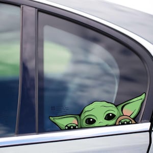 Puede incluir: Una calcomanía de ventana de coche de Baby Yoda verde y beige. La calcomanía se asoma por detrás de la ventana del coche. La calcomanía es una imagen de dibujos animados del personaje de la franquicia Star Wars.