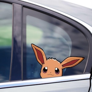 Puede incluir: Una pegatina para ventana de coche con el personaje de dibujos animados Eevee, con orejas grandes y una expresión amigable. La pegatina es marrón con detalles rojos.