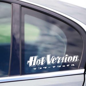 Może przedstawiać: Biała naklejka winylowa z napisem "Hot Version" w stylizowanej czcionce, z japońskimi znakami poniżej. Naklejka jest naklejona na szybie srebrnego samochodu.