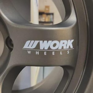 May include: Close-up of a gray alloy wheel with the white text "WORK WHEELS" prominently displayed. The wheel has a multi-spoke design and a metallic finish, suggesting a focus on automotive performance and style.