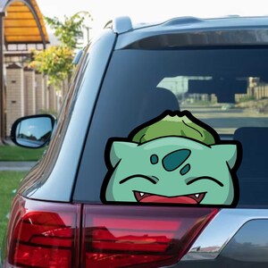 Puede incluir: Una pegatina de dibujos animados verde y negra de un personaje de Bulbasaur está adherida a la ventana trasera de un coche. La pegatina presenta un Bulbasaur sonriente con su característico bulbo en la espalda.