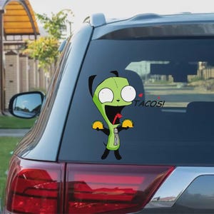 Könnte beinhalten: Ein grüner Cartoon-Charakter-Aufkleber auf einer Autoscheibe, der zwei Tacos hält und daneben das Wort "TACOS!" hat. Die Figur hat große weiße Augen und einen weit geöffneten Mund. Der Aufkleber befindet sich auf der Heckscheibe eines grauen Autos.