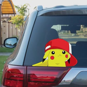 Könnte beinhalten: Ein gelber Pikachu-Aufkleber mit einer rot-weißen Baseballkappe ist auf der Heckscheibe eines Autos angebracht. Der Aufkleber zeigt den Pokémon-Charakter mit roten Wangen und schwarzen Augen.
