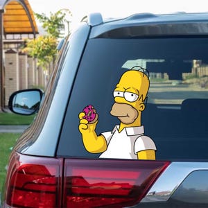 May include: A yellow Homer Simpson sticker, holding a pink donut with sprinkles, on the rear window of a grey car. Homer is wearing a white shirt. The cartoon image is a fun car accessory.