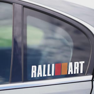 Peut inclure: Autocollant blanc "RALLI ART" avec un motif de rayures rouges, orange et jaunes sur une vitre de voiture. L'autocollant rectangulaire est en vinyle.