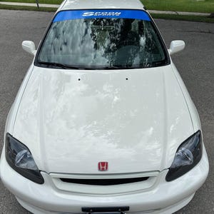 May include: A white saloon car featuring a red Honda emblem on the bonnet, dark-tinted headlights, and a blue windscreen banner with the text Spoon Sports. The vehicle includes a rear roof spoiler and a number plate displaying the text JDM JET.