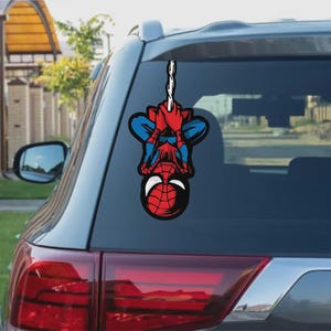 Könnte beinhalten: Ein roter, blauer und schwarzer Spider-Man-Aufkleber ist an der Heckscheibe eines Fahrzeugs angebracht. Der Aufkleber zeigt Spider-Man, der kopfüber an einem weißen Netz hängt. Das Rücklicht des Autos ist darunter sichtbar.