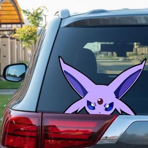 Peut inclure: Un autocollant de voiture Espeon violet et noir est apposé sur la vitre arrière d'une voiture grise. L'autocollant représente le visage du personnage Espeon, avec ses grandes oreilles et sa couleur violette distinctive.