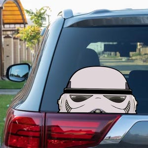 Op de afbeelding: Een wit en zwart Stormtrooper helm sticker op de achterruit van een voertuig. De sticker is een cartoon-stijl illustratie van het iconische Star Wars karakter. Het achterlicht van de auto is rood.
