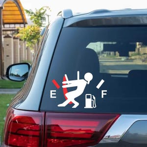 Könnte beinhalten: Weißer Autoaufkleber aus Vinyl, der eine Tankanzeige mit einer Person zeigt, die rennt, um den Tank zu füllen. Die Anzeige zeigt einen niedrigen Kraftstoffstand an, mit dem Buchstaben "E" für leer. Der Aufkleber befindet sich auf der Heckscheibe eines grauen Autos.