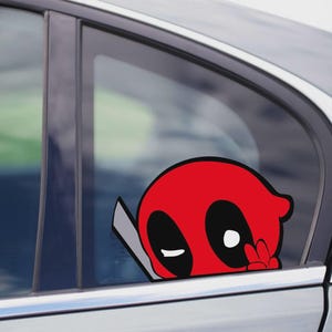 Może przedstawiać: Czerwono-czarna naklejka na samochód Deadpool jest przymocowana do szyby samochodu. Naklejka przedstawia głowę Deadpoola z charakterystycznymi czarnymi oczami i znakiem pokoju. To zabawny i unikalny sposób na spersonalizowanie pojazdu.