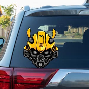 Könnte beinhalten: Ein gelb-schwarzer Cartoon-Aufkleber eines Robotergesichts ist auf der Heckscheibe eines Autos angebracht. Das Design weist ein detailliertes Design mit einem metallgrauen unteren Gesicht und leuchtend gelben Akzenten auf.