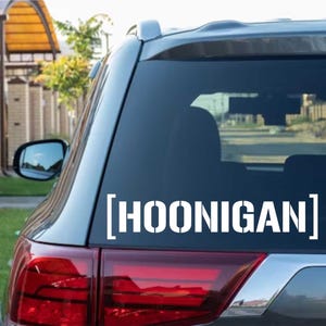 May include: White vinyl decal with the word "HOONIGAN" in a stencil font, displayed on the rear window of a gray SUV. The decal is centered and clearly visible against the dark glass.
