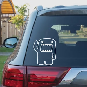 May include: White vinyl decal of a cartoon character with a large mouth and waving arm, applied to the rear window of a gray car. The character has a rectangular body and is outlined in white.