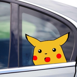 May include: A yellow Pikachu car decal peeking out of a car window. The decal features Pikachu's face with black eyes, red cheeks, and an open red mouth, creating a surprised expression. The car is silver.