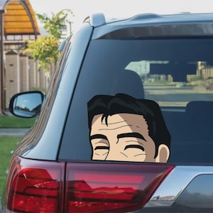 Könnte beinhalten: Ein Autoaufkleber mit einer Cartoon-Illustration eines Mannes mit geschlossenen Augen und schwarzen Haaren. Der Aufkleber befindet sich auf der Heckscheibe eines grauen Autos, mit dem Rücklicht darunter.
