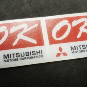 May include: Two rectangular stickers featuring the Mitsubishi Motors Corporation logo. Each sticker has a red background with the white letters "OK" and the Mitsubishi logo. The text "MITSUBISHI MOTORS CORPORATION" is printed below the logo.