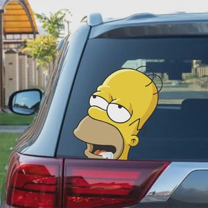 May include: A yellow Homer Simpson car sticker with a surprised expression is applied to the rear window of a grey car. The sticker features Homer's face with his mouth open and eyes wide.