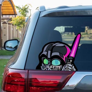 Peut inclure: Autocollant de voiture Darth Vader noir et gris avec un sabre laser rose. L'autocollant est appliqué sur la vitre arrière d'une voiture, montrant la tête et l'arme du personnage. Le design est de style cartoon.