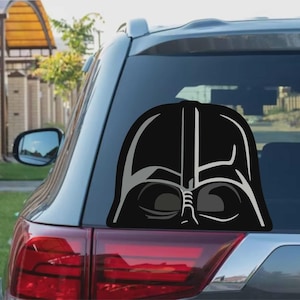 Op de afbeelding: Een zwarte en grijze Darth Vader helm sticker is bevestigd op de achterruit van een grijze auto. De sticker toont het iconische helmontwerp uit de Star Wars-franchise, met gedetailleerde gezichtskenmerken en een glanzende afwerking.