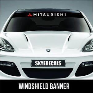 Op de afbeelding: Een witte auto met een zwarte voorruitbanner met het woord "MITSUBISHI" in witte tekst en een rood logo. De nummerplaat toont "SKYEDECALS" en de onderste banner "WINDSHIELD BANNER".