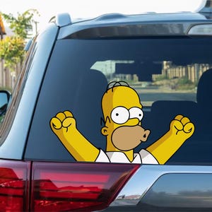 May include: A yellow Homer Simpson car sticker with his arms raised in a celebratory pose. The sticker is on the rear window of a grey SUV, with the taillight visible below. The cartoon character is wearing a white shirt.