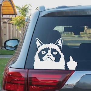 May include: White vinyl decal of a grumpy cat with a raised middle finger, applied to the rear window of a gray car. The cat's face is detailed with black markings, conveying a stern expression. The decal is a humorous accessory.