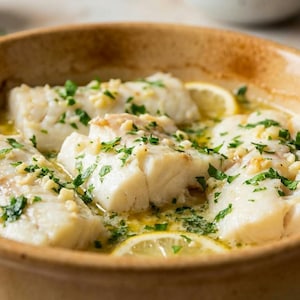 May include: A close-up shot of baked fish fillets in a light brown ceramic dish. The fish is topped with chopped parsley and garlic, and lemon slices are visible. The dish is filled with a buttery sauce.