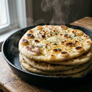 May include: A stack of freshly baked naan bread in a cast iron skillet. The naan is golden brown with dark spots, and steam rises from the warm bread. The skillet sits on a rustic wooden table near a window.