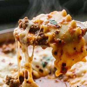 Puede incluir: Primer plano de una cuchara de sopa de lasaña. La cuchara contiene un trozo grande de pasta, carne picada y queso derretido. La sopa está en un cuenco blanco, con vapor saliendo de la comida caliente.