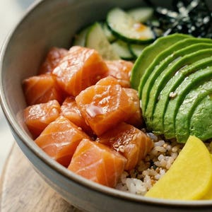 May include: A close-up of a poke bowl filled with salmon cubes, sliced avocado, cucumber, edamame, and pickled ginger. The bowl sits on a wooden surface. The dish is a colorful and healthy meal.