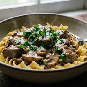 Könnte beinhalten: Eine Schüssel mit Beef Stroganoff mit Eiernudeln, Pilzen und Petersilie. Das Gericht wird in einer gesprenkelten Keramikschüssel mit einer Gabel serviert. Dampf steigt von dem warmen Essen auf, das auf einem rustikalen Holztisch steht.