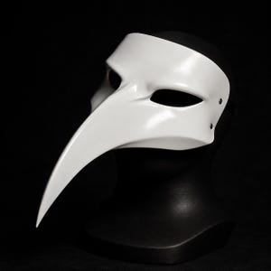 May include: A smooth, white plague doctor mask featuring a long, curved beak and almond-shaped eye openings. The mask is constructed with a solid, glossy finish and includes small metal rivets on the side where the strap attaches to the frame.