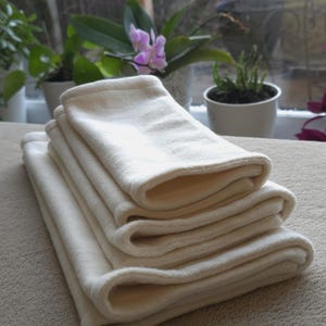 Op de afbeelding: Een stapel van vier gevouwen, crèmekleurige stoffen luiers. De luiers zijn gemaakt van een zacht, absorberend materiaal en zijn netjes op een lichtbeige oppervlak geplaatst. De afbeelding is goed belicht, waardoor de textuur en plooien van de stof zichtbaar zijn.
