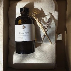 Puede incluir: Una botella de vidrio ámbar oscuro de aceite de ricino, 250 ml, con una etiqueta blanca, se encuentra en una caja de cartón con una tela blanca y ramitas de trigo y lavanda. La etiqueta dice "Castor Oil" y "Natural | Clean | Honest".