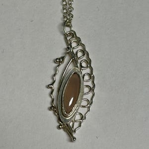 May include: A silver pendant necklace featuring a marquise-shaped, smooth, light brown stone set in a decorative, open-work silver frame with wire-looped edges. The pendant hangs from a delicate silver chain with small, circular metal links.