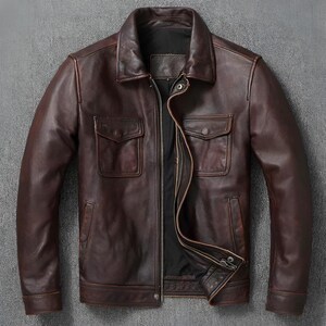 Könnte beinhalten: Eine dunkelbraune Lederjacke mit klassischem Kragen, durchgehendem Frontreißverschluss und zwei Brusttaschen mit geknöpften Klappen. Die Jacke hat lange Ärmel mit Manschetten, eine glatte Oberfläche und ein schwarzes Innenfutter an der Öffnung.