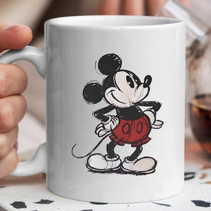 May include: White ceramic mug with a sketched illustration of Mickey Mouse. The cartoon character is shown in his classic outfit: red shorts, white gloves, and large white shoes. The mug has a curved handle and is held by a person.