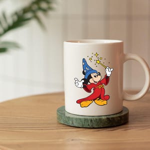 May include: A white ceramic mug with a colourful illustration of Mickey Mouse as a sorcerer, holding a wand with star accents. The mug is on a green marble coaster, placed on a wooden surface.