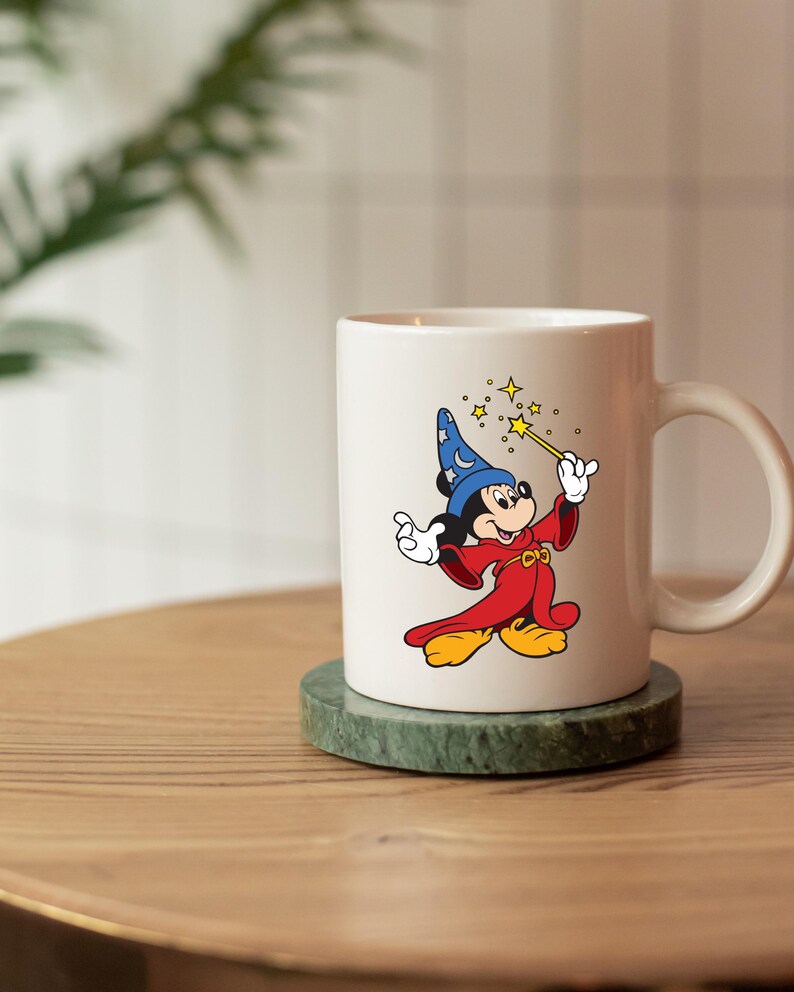 May include: A white ceramic mug with a colourful illustration of Mickey Mouse as a sorcerer, holding a wand with star accents. The mug is on a green marble coaster, placed on a wooden surface.