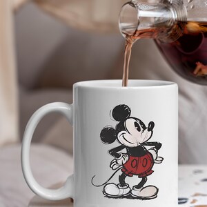 May include: White ceramic mug with a sketch-style Mickey Mouse illustration. The mug has a curved handle and is being filled with a dark liquid from a glass teapot. A classic Disney design.