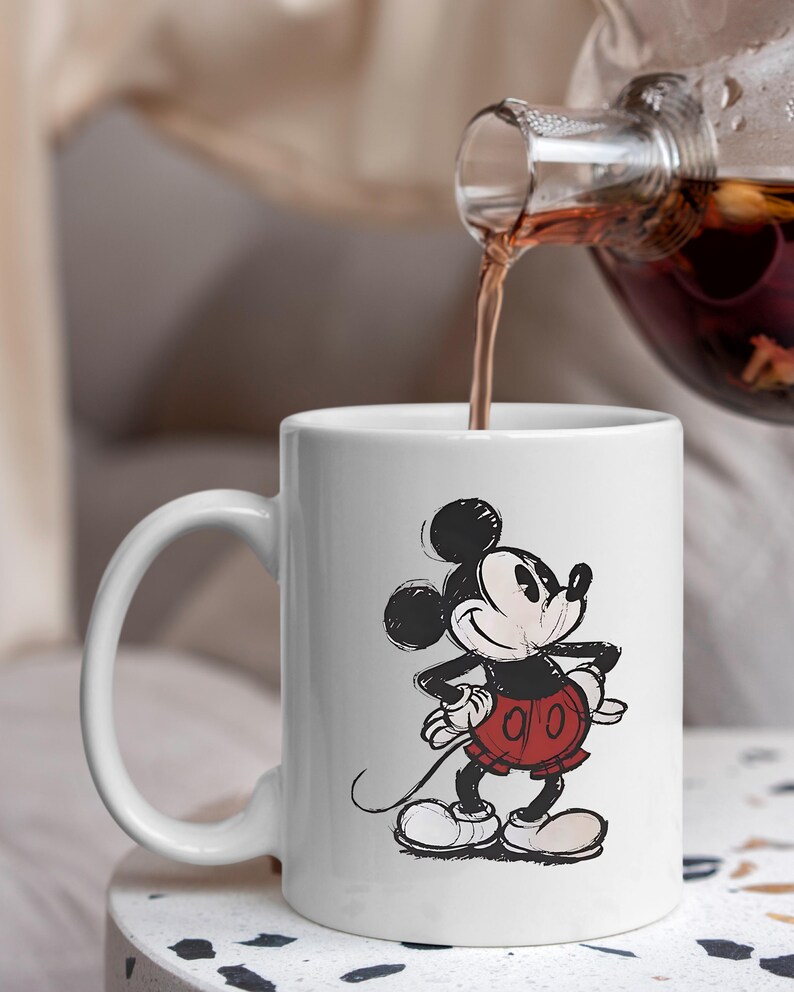 May include: White ceramic mug with a sketch-style Mickey Mouse illustration. The mug has a curved handle and is being filled with a dark liquid from a glass teapot. A classic Disney design.